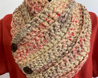 May include: A chunky, hand-crocheted infinity scarf in a blend of beige, red, and purple yarns. The scarf features a button closure with three dark brown wooden buttons. The thick yarn creates a textured, warm appearance.