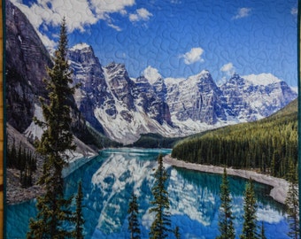 Può includere: Un arazzo trapuntato raffigurante un paesaggio montano riflesso in un lago turchese. L'immagine mostra cime innevate, alberi sempreverdi e un cielo blu con nuvole. La trapunta ha un bordo verde.