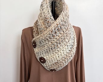 Può includere: Una sciarpa a collo spessa, lavorata a mano all'uncinetto, nei toni neutri del beige, grigio e marrone. La sciarpa presenta una chiusura con bottoni con tre grandi bottoni in legno marrone scuro. Il filato spesso crea un accessorio caldo e strutturato.