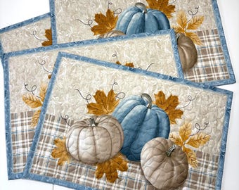 Può includere: Un set di tovagliette rettangolari trapuntate con una stampa stagionale di zucche blu e beige circondate da foglie d'acero color marrone dorato. Lo sfondo presenta un motivo a volute color crema e un bordo a quadri marrone.