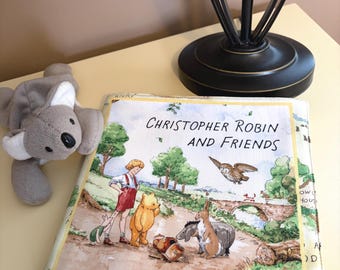 Può includere: Un libro di stoffa intitolato "Christopher Robin and Friends" con illustrazioni di Winnie the Pooh e dei suoi amici. Un peluche di koala grigio è appoggiato accanto al libro. È visibile anche una lampada nera con base decorativa.