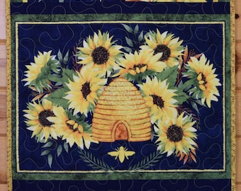 Può includere: Una trapunta con sfondo blu e un motivo di girasoli e un alveare. I girasoli sono gialli con centri marroni e foglie verdi. L'alveare è giallo e marrone. La trapunta è incorniciata da un bordo giallo.