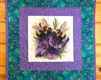 May include: A square quilted wall hanging featuring a central image of two bumblebees amidst purple flowers. The artwork is framed by a purple border with a swirling pattern, and a green and blue floral patterned border. The background is a wooden wall.