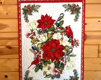 May include: A rectangular quilted wall hanging featuring a central arrangement of red poinsettias, white magnolia flowers, pine cones, and holly berries. Four birds, including two red cardinals and two chickadees, are perched among the foliage.