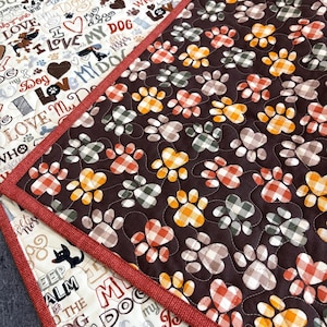 May include: A quilted fabric mat featuring two distinct patterns. One side displays a dark brown background with repeating paw prints in orange, yellow, and grey plaid. The other side features a cream background with text and dog-themed graphics.