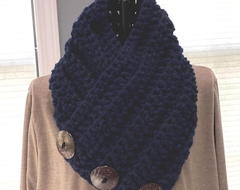 May include: A navy blue crocheted cowl scarf with three large, round, wooden buttons. The scarf is made of a thick, textured yarn and is displayed on a mannequin. The buttons are a light brown color.