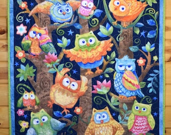 May include: A rectangular quilt featuring a dark blue background with a forest scene of brown tree trunks, green vines, and colorful flowers. Nine stylized owls in various colors including orange, blue, green, and pink are perched on branches.