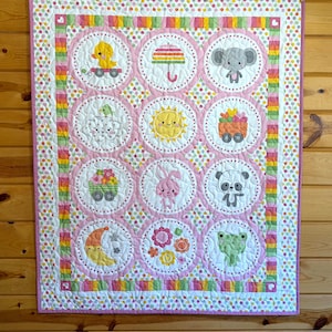May include: A handmade baby quilt with a repeating pattern of circular designs featuring cute animals and objects. The quilt has a pink and white border with colorful polka dots and is hanging on a wooden wall.