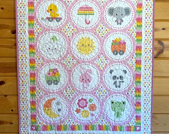 May include: A handmade baby quilt with a repeating pattern of circular designs featuring cute animals and objects. The quilt has a pink and white border with colorful polka dots and is hanging on a wooden wall.