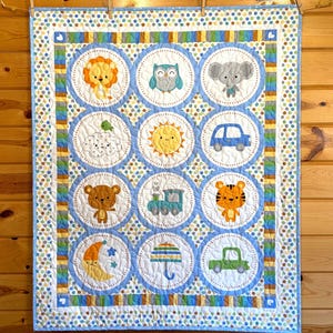 May include: A baby quilt with a repeating pattern of circular designs featuring various animals, vehicles, and celestial bodies. The quilt has a blue and white color scheme with accents of yellow, orange, and green. The quilt is hanging on a wooden wall.