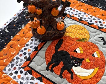 May include: A quilted fabric mat featuring a black cat and a large orange jack-o'-lantern, topped with a brown decorative tree sculpture adorned with several small orange pumpkin ornaments with carved facial expressions.
