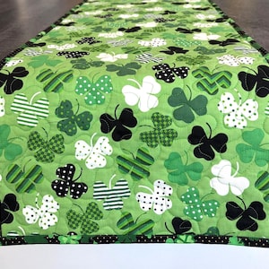 May include: A green quilted table runner featuring a pattern of shamrocks in various shades of green, white, and black. The shamrocks have different designs, including polka dots and stripes. The border is black with white polka dots.