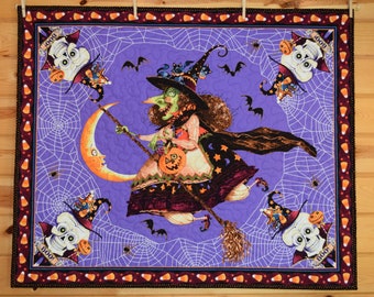 Può includere: Una trapunta a tema Halloween con una strega su una scopa su uno sfondo viola con un motivo a ragnatela. La trapunta ha un bordo con motivi di caramelle e teschi. La strega tiene in mano una zucca.