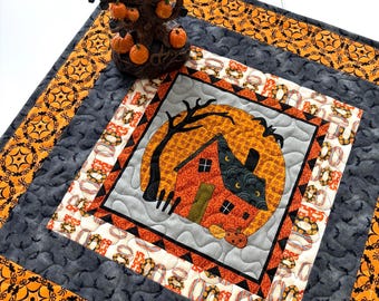 Può includere: Un runner da tavola trapuntato quadrato con un blocco centrale raffigurante una luna arancione e una casa, circondato da bordi con ragnatele, pipistrelli e zucche. Un albero decorativo marrone con piccole zucche arancioni è posto sopra.
