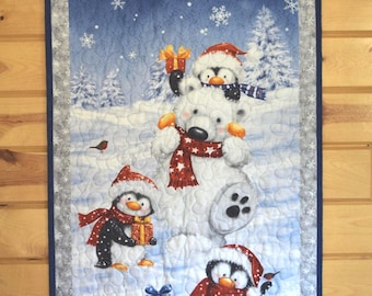 May include: A rectangular fabric wall hanging featuring a winter scene with a white polar bear and three penguins wearing red hats and scarves. The scene includes falling snowflakes, pine trees, and wrapped gifts on a snowy ground with a gray border.