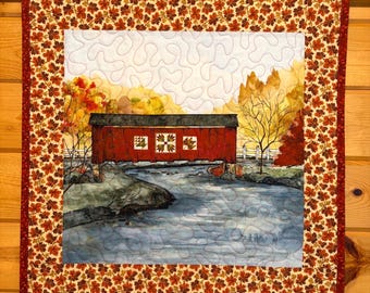 May include: A square fabric wall hanging featuring a printed scene of a red covered bridge over a river, surrounded by autumn trees. The central panel is framed by a border of small orange, yellow, and brown maple leaves on a dark red background.
