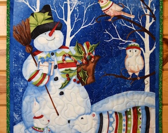 May include: A rectangular fabric wall hanging featuring a winter scene with a snowman holding a broom and a gift, two white dogs wearing striped sweaters, a bird, and an owl in a snowy forest with blue sky and white snowflakes, finished with a green border.
