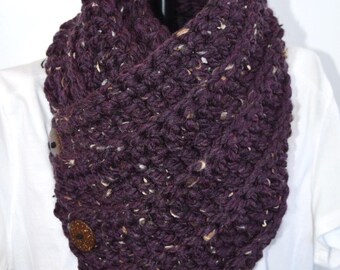 May include: A chunky knit cowl scarf in a deep plum purple color with flecks of tan and cream throughout the yarn. The scarf features three round, dark brown textured buttons arranged vertically along the side edge of the thick fabric.