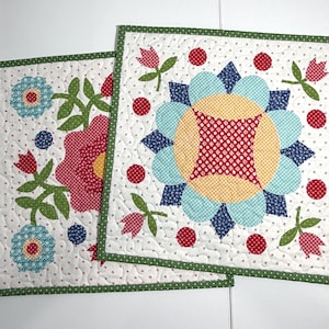 May include: Two square quilted fabric mats featuring a white background with small red floral patterns. The designs include appliqued flowers in shades of red, yellow, and light blue, accented by green stems and leaves, all finished with a green border.