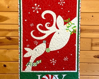 May include: A Christmas-themed wall hanging featuring a red background with white reindeer and snowflakes. The word "JOY" is displayed at the bottom, with a peppermint candy design in the letter "O". The border is green with a white and red zigzag pattern.