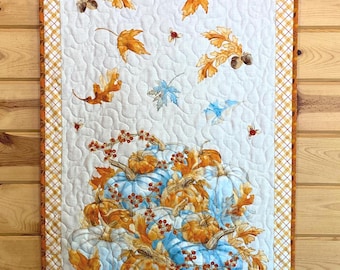 Può includere: Un arazzo trapuntato a tema raccolto autunnale. Il design include zucche azzurre, foglie arancioni e bacche su sfondo bianco. Il bordo presenta un motivo a quadri arancione e marrone.