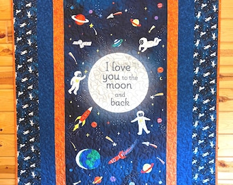May include: A quilted wall hanging with a space theme. The central design features a dark blue background with planets, rockets, astronauts, and the text "I love you to the moon and back." The quilt is bordered with blue and orange fabrics, and a patterned edge.