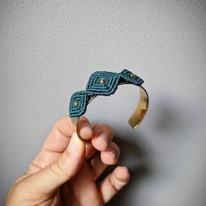 May include: A gold metal bangle bracelet with a teal blue macrame design. The macrame design features a series of connected squares with gold beads in the center of each square.