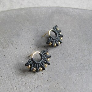 May include: A pair of small, black and gold earrings. The earrings are made of black thread and have a small gold circle in the centre. The gold circle is surrounded by a ring of small gold beads.