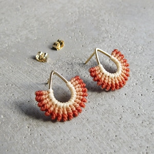 May include: A pair of teardrop-shaped earrings with a graduated ombre design in shades of coral and cream. The earrings are made with a macrame technique and have gold-toned posts.