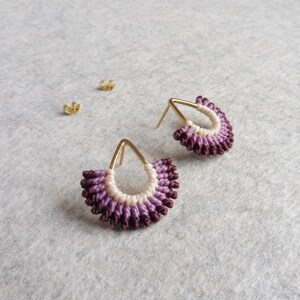 Peut inclure: Une paire de boucles d'oreilles en or en forme de goutte. Les boucles d'oreilles sont faites d'une ficelle violette et blanche tissée en forme d'éventail.