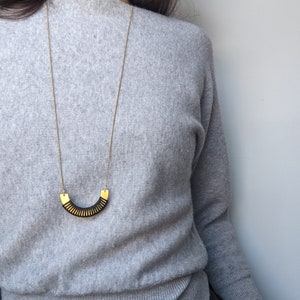 May include: A gold chain necklace with a black and gold geometric pendant. The pendant is shaped like a crescent moon and has a textured surface.