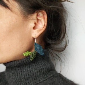 Puede incluir: Un par de pendientes en forma de hoja verde y azul hechos con nudos de macramé. Los pendientes cuelgan de ganchos plateados.