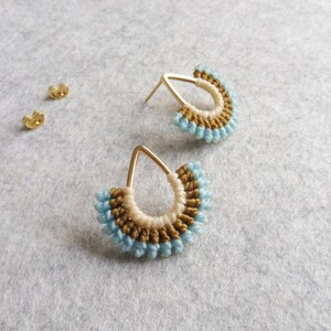 Peut inclure: Une paire de boucles d'oreilles en forme de goutte d'eau dorées avec un motif perlé. Les perles sont dans des tons de bleu, blanc et or.