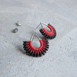 May include: A pair of handmade earrings with a distinctive design. The earrings have a silver hoop with a fan-shaped pattern in red and black. The earrings are made using a macrame technique and have a textured appearance. Earring backs are also visible.