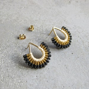 May include: A pair of gold hoop earrings with black, white, and gold thread wrapped around them. The earrings are teardrop shaped and have a delicate design.