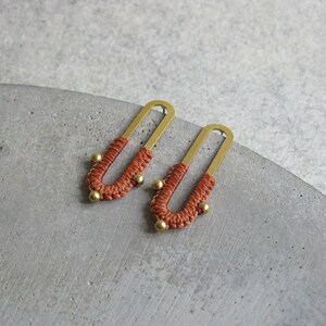 May include: A pair of earrings with a gold metal frame and a woven orange cord. The cord is wrapped around the frame in a U-shape and has small gold beads at the ends.