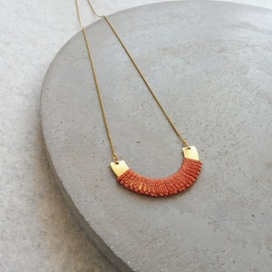 May include: A gold chain necklace with a red woven pendant. The pendant is shaped like a crescent moon.