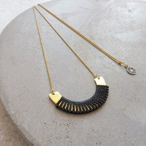 May include: A gold chain necklace with a black woven pendant. The pendant is shaped like a crescent moon and has a gold trim.