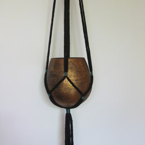 May include: Black macrame plant hanger with a copper-colored pot. The hanger has a tassel at the bottom and is made with a dark green accent color.