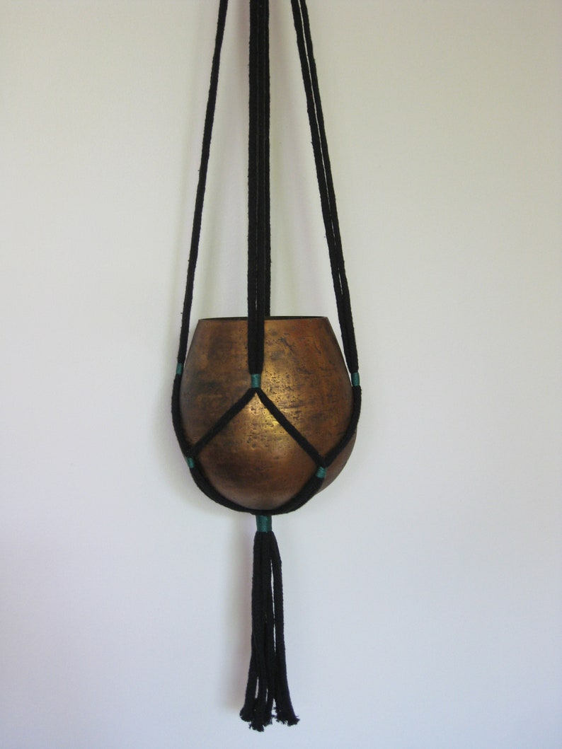 May include: Black macrame plant hanger with a copper-colored pot. The hanger has a tassel at the bottom and is made with a dark green accent color.