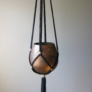 May include: Black macrame plant hanger with a copper pot. The hanger has wooden beads and a tassel.