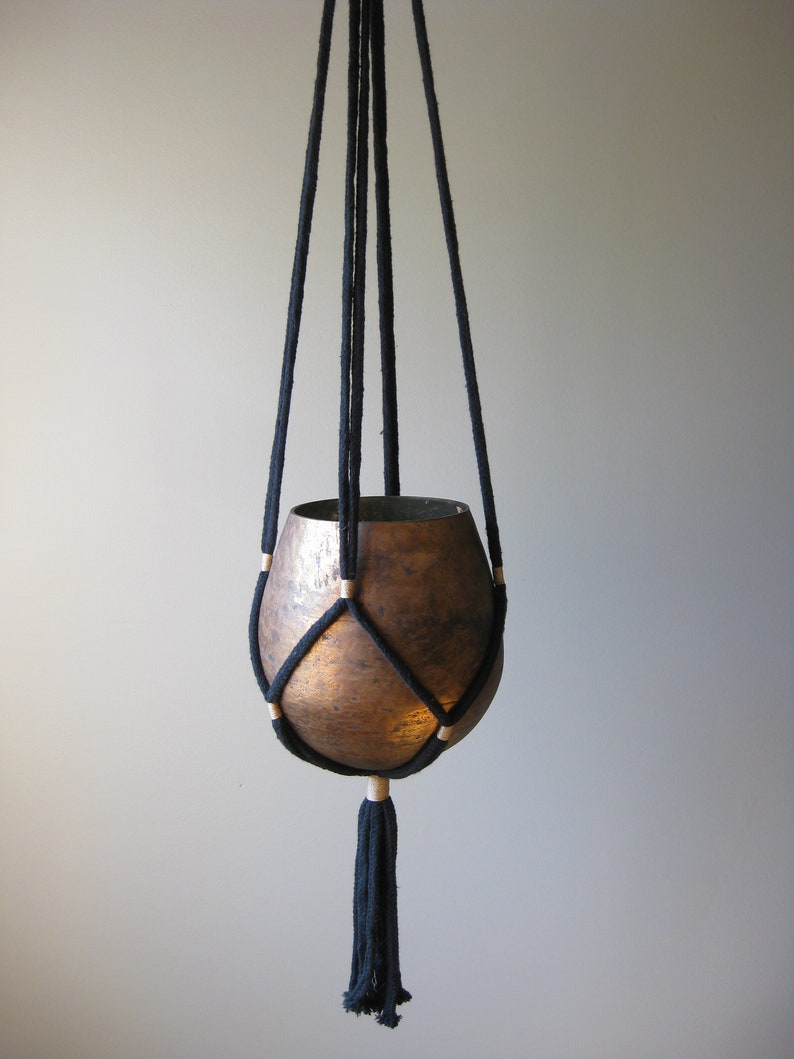 May include: Black macrame plant hanger with a copper pot. The hanger has wooden beads and a tassel.