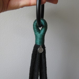 May include: A black macrame plant hanger with a green rope detail and a silver charm. The hanger is attached to a black ring.