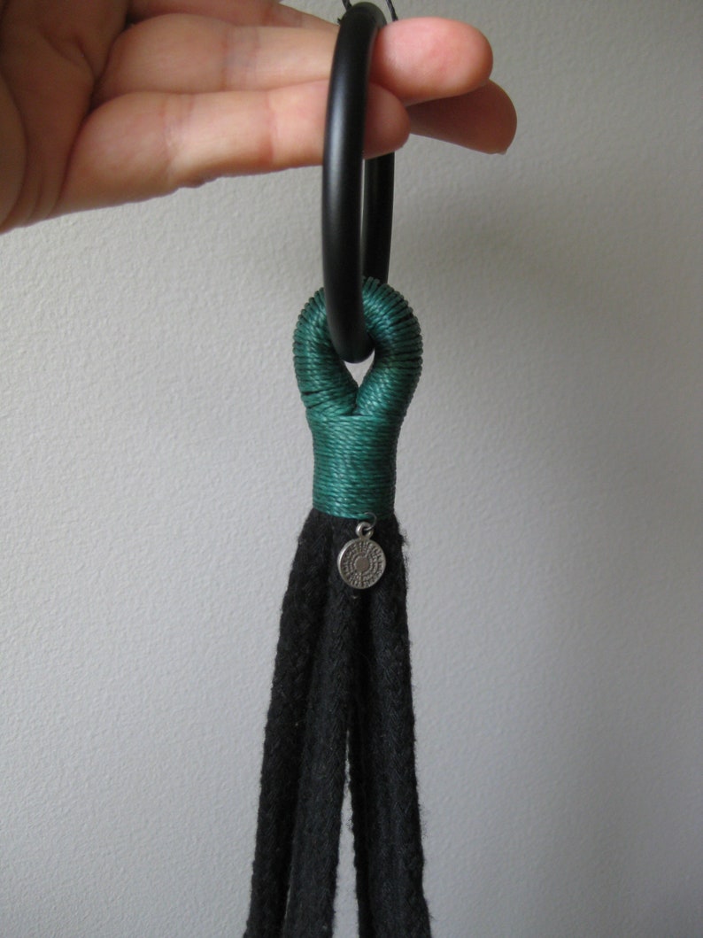 May include: A black macrame plant hanger with a green rope detail and a silver charm. The hanger is attached to a black ring.