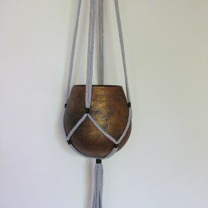 May include: A gray macrame plant hanger with a brown ceramic pot. The hanger has a tassel at the bottom.