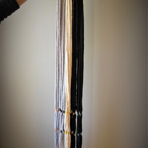 May include: Five macrame plant hangers with different colored cords, including gray, white, and black. Each hanger has a black metal ring at the top and a tassel at the bottom.