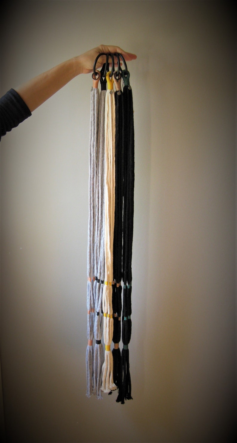 May include: Five macrame plant hangers with different colored cords, including gray, white, and black. Each hanger has a black metal ring at the top and a tassel at the bottom.