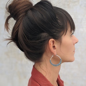May include: A woman with dark brown hair in a bun is wearing large hoop earrings with blue and gold beads. The earrings are silver and have a simple, elegant design.