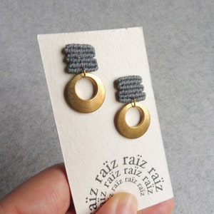 Peut inclure: Une paire de boucles d'oreilles grises et dorées. Les boucles d'oreilles sont faites d'un tissu gris tissé et d'un cercle doré avec un trou au centre. Les boucles d'oreilles sont présentées sur une carte blanche avec le mot "raíz" répété plusieurs fois.