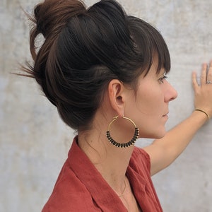 May include: A woman with dark brown hair in a bun is wearing large gold hoop earrings with black beads. She is wearing a rust coloured shirt.